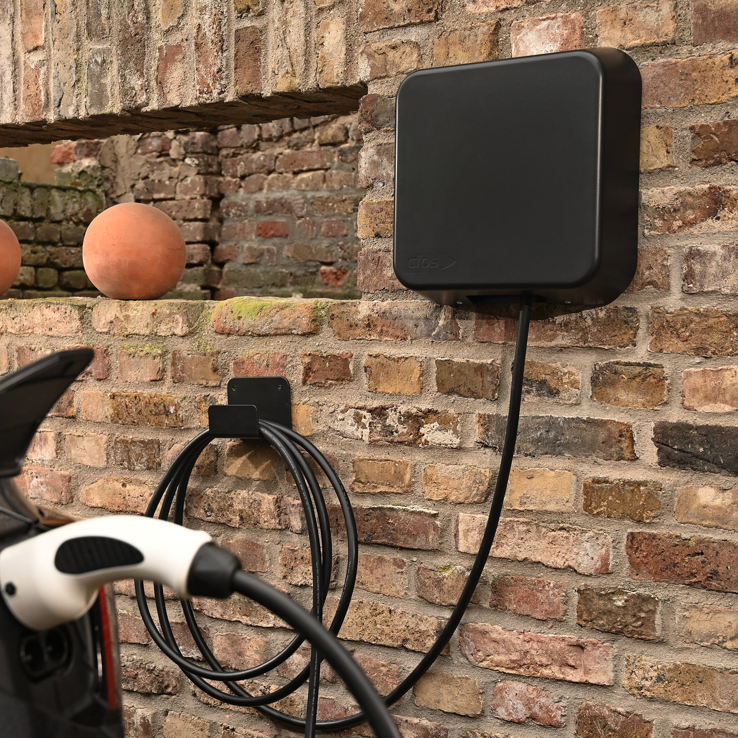 The picture is showing a cfos emobility wallbox ng mounted on a brickwall. The attached type two charging plug is charging a car. The cable has a length of seven meters and is rolled up on the cable-holder that is included in the product.
